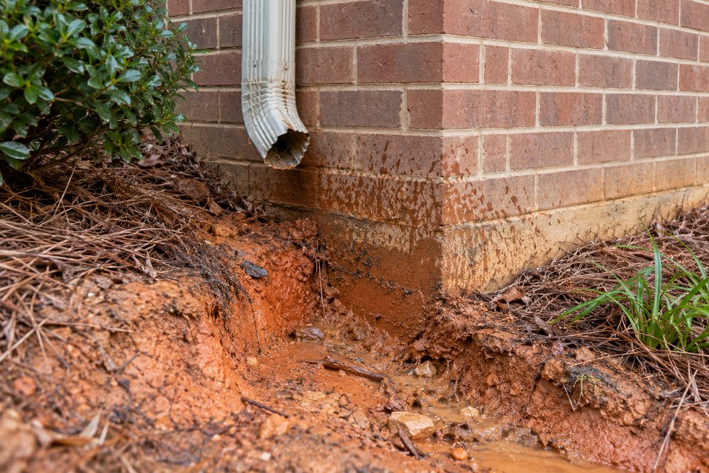Washed-out red clay and muddy splash marks at a brick foundation corner below a short broken downspout